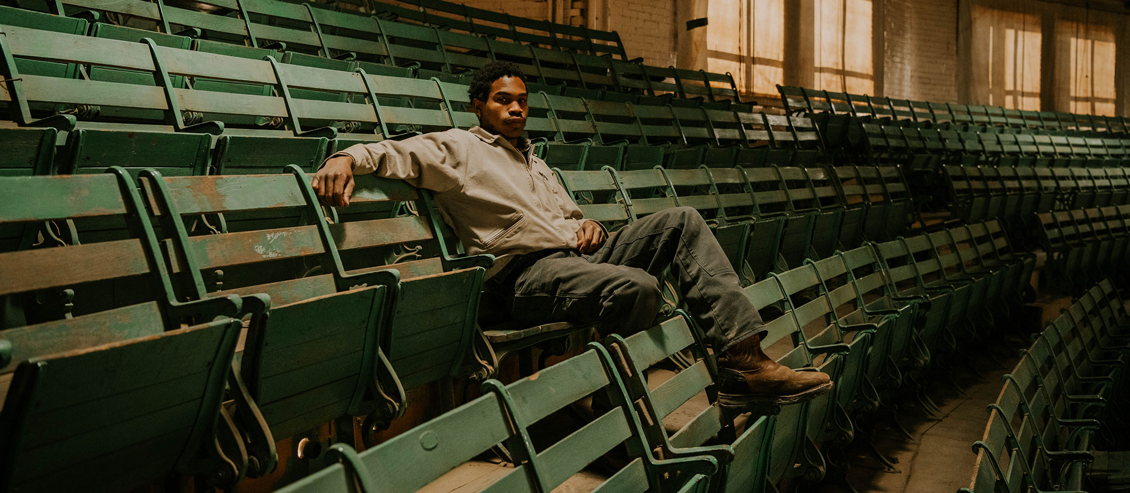 A man sits in the stands at the cattle sale barn, posing and wearing workwear.